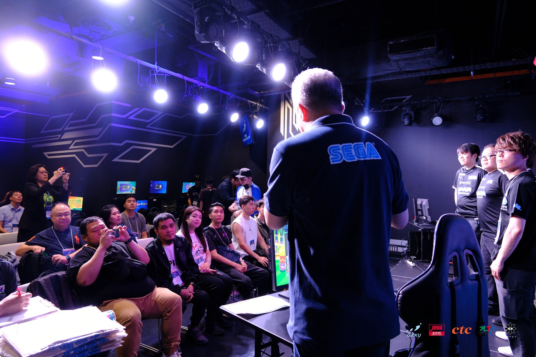 A SEGA coach addressing a seated international cohort inside a darkened Tokyo esports lab